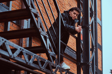 Fototapeta na wymiar A fashionable guy leaning on a guardrail of stairs outside the building with the industrial exterior.