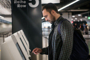Young man with backpack touching interactive display using self service machine, doing...