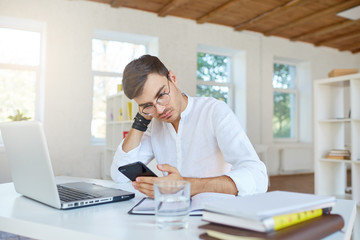 Indoor shot of concentrated tired young businessman wears white shirt and glasses using laptop and cell phone sitting and working at the table in office