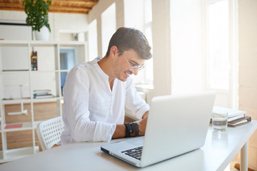 Indoor shot of cheerful handsome young businessman wears white shirt and spectacles using laptop and working at the table in office