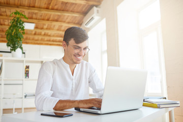 Portrait of cheeful confident young businessman wears white shirt and spectacles using laptop and smartphone working at the table in office and laughing