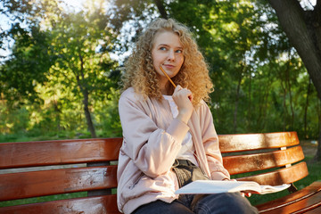 A young beautiful girl blonde sits on a park bench, with headphones, notes, a notepad, looks away,...