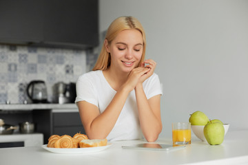 Obraz na płótnie Canvas Young blonde girl sits at the kitchen table in the morning, smiling, looks at tablet black screen, on the table -a glass of juice, apples, buns, dressed in a domestic white T-shirt, kitchen background