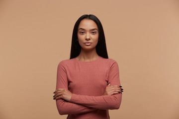 Obraz premium Portrait of calm, confident, attractive young mixed race brunette girl stands with arms crossed, dressed casual, on beige background
