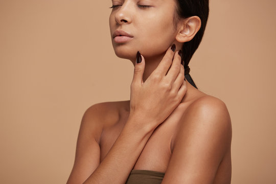 Portrait Of An Attractive Mixed Race Girl, Stands Sideways, Demonstrates Beautiful Skin After The Procedure, Tenderly Touches Her Neck, Eyes Closed, Feels Calmness And Pleasure, Over Beige Background