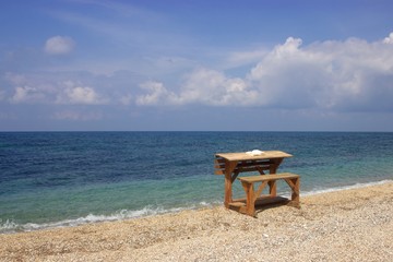 Wooden desk on a beach with an open book on it and blue sea and clouds at the background 