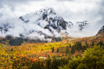 A Cabin in a Forest with Fall Colored Leaves Below Cloud Covered Mountains With Snow