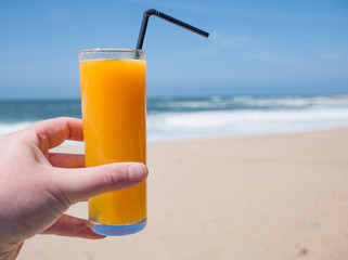 Male hand holding glass of freshly squeezed orange juice with a straw and blue ocean and sandy beach behind. Shallow depth of field