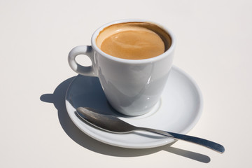 Cup of hot fresh coffee in a white cup with white saucer and a spoon on an off-white table viewed from the front.