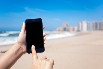 Woman's hands holding and using smartphone on beach with deep blue sky and golden sand. The phone has a black screen
