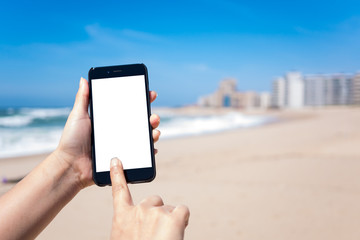 Woman's hands holding and using smartphone on beach with deep blue sky and golden sand. The phone has a keyed blank white screen