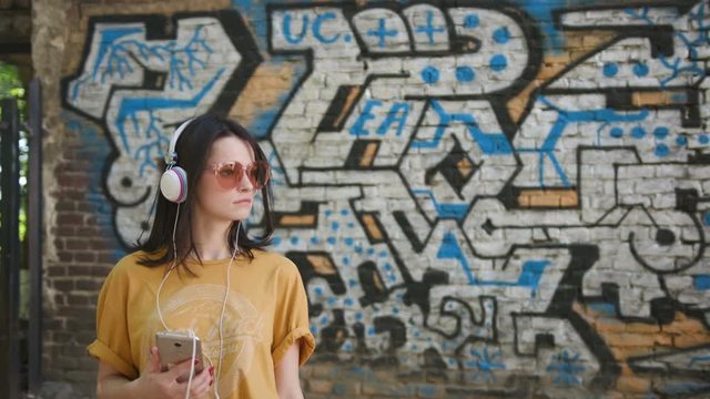 Nice teenage girl wearing headphones listening to music. Student walking through the sports ground. Street art in background. Hip-hop concept. Painted wall. Creative art.