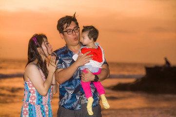 young happy and beautiful Asian Japanese couple holding baby girl daughter walking on sunset beach enjoying together romantic summer holidays trip