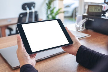 Mockup image of hands holding and using black tablet pc with blank white desktop screen with notebook on wooden table in office