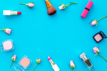 Rose, pink decorative cosmetics frame. Lipstick, bulk, eyeshadow and small rose flowers on blue background top view copy space