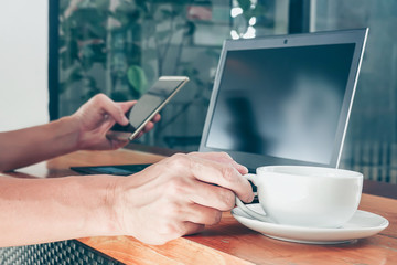 Mockup image of man's hands holding mobile phone with blank black screen and laptop and a coffee cup on wooden table.