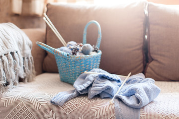 Knitting in cozy home interior. Winter or Christmas mood, atmosphere. Knitted clothes, blue wicker basket with yarn, woolen balls, needles on sofa in warm sunlight. Concept of woman's hobby, leisure.
