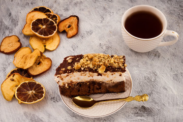 A sliced piece of cake on a plate. A layer of white meringue, chocolate dough, chopped nuts. Dry fruit slices. White mug with tea. Light background.