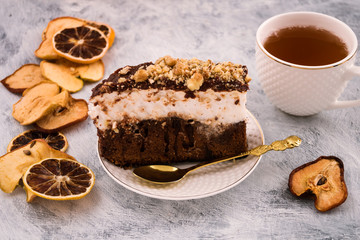 Sweet dessert on the plate. Piece of chocolate cake. Layer of white meringue. Grated walnut. Dried fruit slices. White mug with a drink. Light background.
