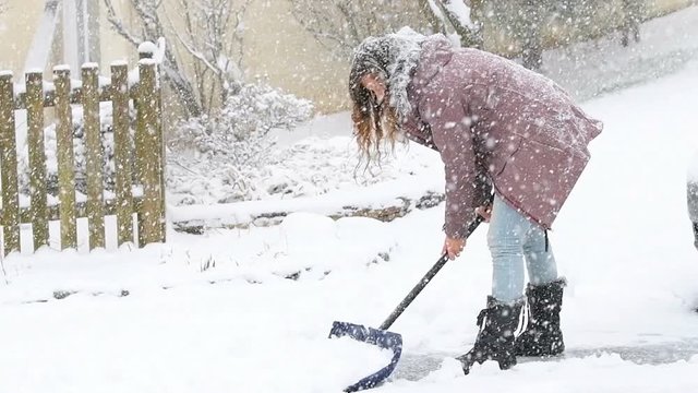 Slow motion of young woman, female in winter coat cleaning, shoveling driveway, street, road from snow in heavy snowing snowstorm with shovel, residential houses