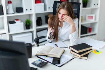 A young girl working in the office with books and documents. The girl took off her glasses and rubs her eyes.