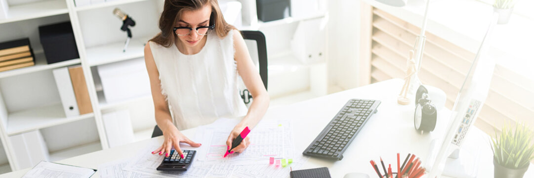 A young girl in the office holds a pink marker in her hand and counts on a calculator.
