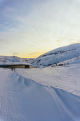 Fototapeta na wymiar two people are walking in the distance along a snowy winter road in the village beyond the Arctic Circle to meet bright sunset light and a small sea bay