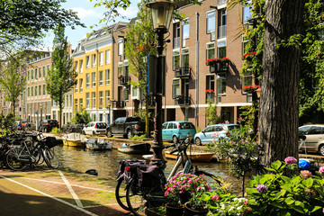 Colourful townhouses over the canal in Amsterdam. Cars and bicycles parked  on the both sides of the canal. Boats moored at the canal bank. Red and pink flowers in the pots, green foliage.