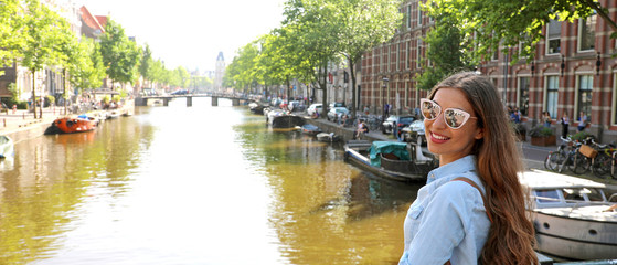 Portrait of traveler girl with sunglasses and backpack enjoying Amsterdam city. Panoramic banner view of young woman looking to the camera on Amsterdam channel, Netherlands, Europe. - obrazy, fototapety, plakaty