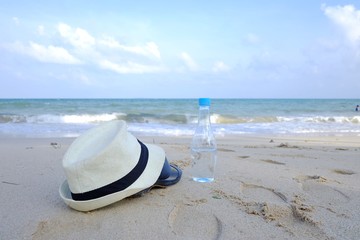 Fototapeta na wymiar White hat with a sandals and a drinking water bottle on the beautiful sea beach in bright day,a horizontal line with blue sky white clouds and seascape for a travelling concept 