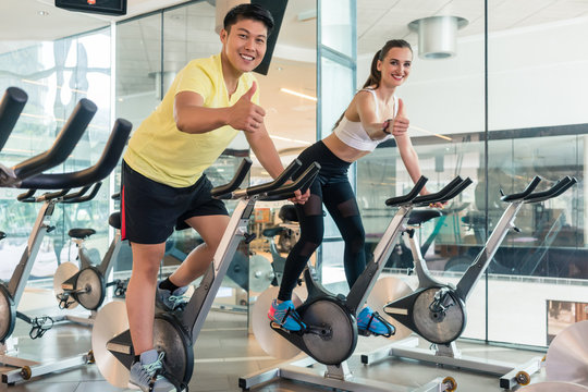 Young cheerful couple showing thumbs up while looking at camera during indoor cycling at the gym