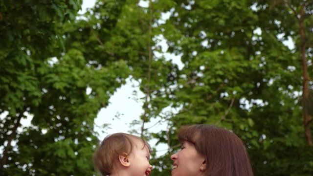 Mom and little daughter are playing in park and laughing. Mom throws happy child to top on background of trees. Slow motion.