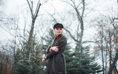 A girl on a background a park with fir-trees is dressed in an overcoat and cap