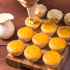 Female confectioner in a blue apron and a plaid red shirt applies cream to cupcakes from a pastry bag