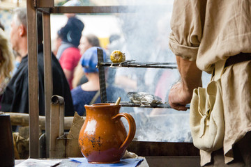 Unidentified man in medieval clothes is preparing barbecue with fish over open fire. Selective focus on the jug.