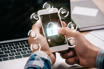 cropped shot of businessman using smartphone with cyber security signs at workplace with laptop