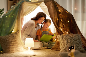 Obraz na płótnie Canvas family, hygge and people concept - happy mother and little daughter reading book in kids tent at night at home