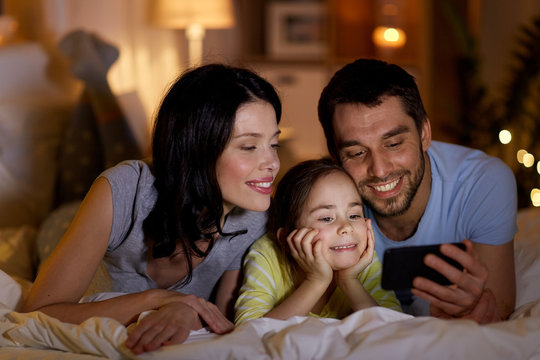 people and family concept - happy mother, father and little daughter with smartphone in bed at night at home