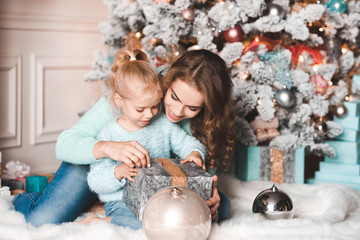 Happy mother and baby daughter open Christmas presents over decorations in room. Winter season.