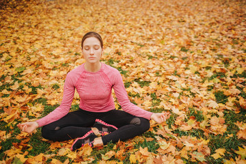 Serious and concentrated young woman sits in lotus position and keeps eyes closed. She is calm and peaceful. Woman sitson ground full of leaves. It is autumn outside.