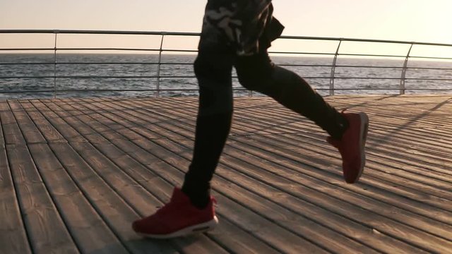 Cropped male legs of healthy sportsman wearing sport leggings and sneakers running along pier at seaside during beautiful morning sunrise