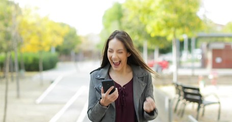 Front view of an excited woman walking towards camera, checking phone and celebrating good news in a park - Powered by Adobe