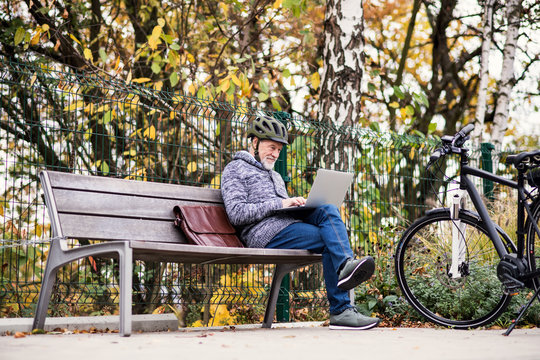 A senior man with electrobike sitting on a bench outdoors in town, using laptop.