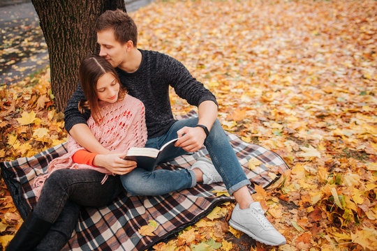 Lovelu picture of young man sitting on blanket with woman . He embrace and kiss her. She looks at book. They hold it together.