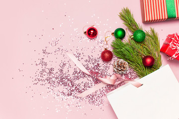 Flat lay top view White gift bag holographic glitter confetti red green christmas balls pine branches gift boxes on pink background. Greeting card Festive holiday decoration. Congratulations New Year