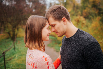 Picture of young man and owman in love. They hold heads close to each other. She smiles. They keep eyes closed. People are in park.