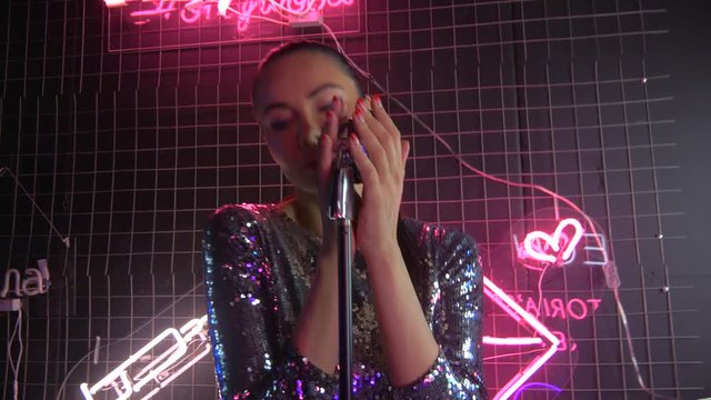 A girl in a gorgeous tantset dress with a microphone. Neon lamps, shiny metal microphone on the stand. A girl in club clothes is dancing and singing into a microphone. 4K video.