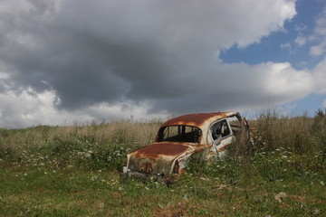 Abandoned old rusty vintage car on hill at summer with clouds at sky inside flowers green plants