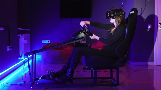 Young woman playing in car drive simulator at VR club, using modern virtual reality headset. Future and video game concept, overall plan