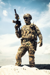 Fototapeta premium Man in military camouflage uniform and mask, equipped tactical ammunition, standing on sand dune with service rifle replica in hands, cloudy sky on background. Airsoft player taking part in war games